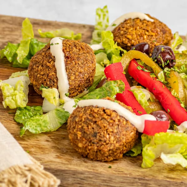 Three falafel balls with white sauce, lettuce, red peppers, and olives on a wooden board.