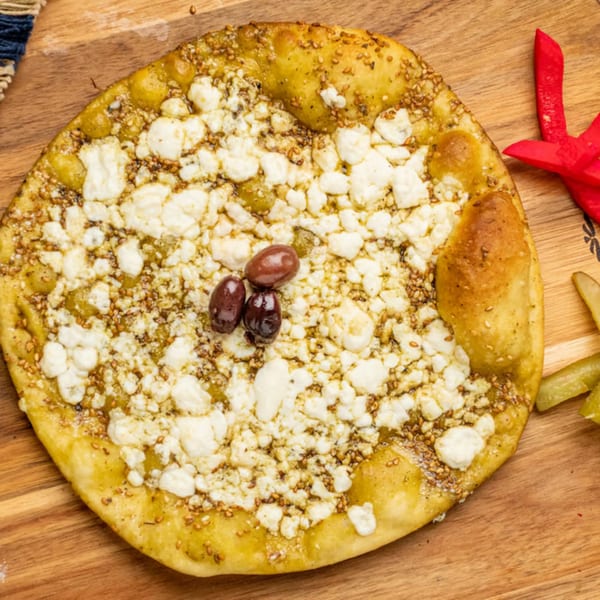 A Mediterranean flatbread topped with white cheese, za'atar, and three dark olives on a wooden board.