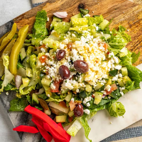 A vibrant Mediterranean salad with lettuce, cucumbers, tomatoes, feta, olives, and pickles on a wooden board.