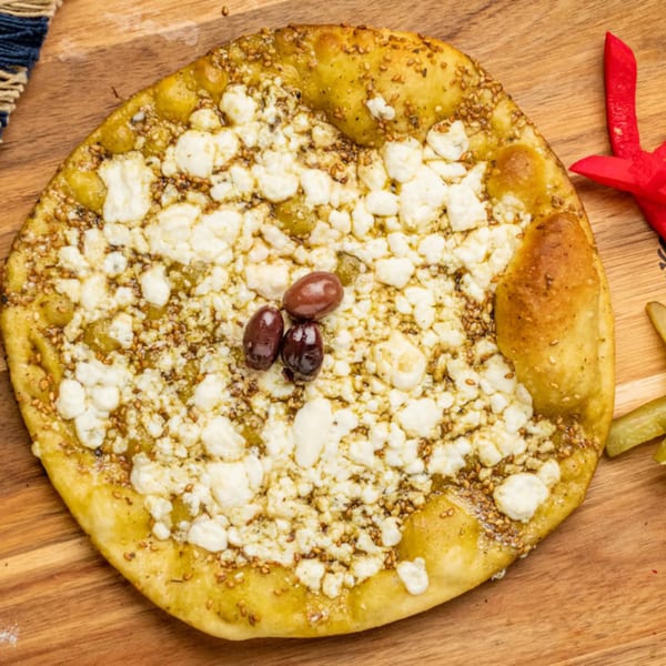 A Mediterranean flatbread topped with white cheese, za'atar, and three dark olives on a wooden board.