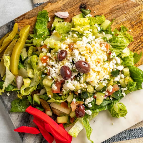 A vibrant Mediterranean salad with lettuce, cucumbers, tomatoes, feta, olives, and pickles on a wooden board.